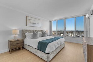 Un dormitorio moderno con una cama grande, una mesita de noche, una lámpara, un cuadro en la pared y vistas a la ciudad a través de amplias ventanas.
