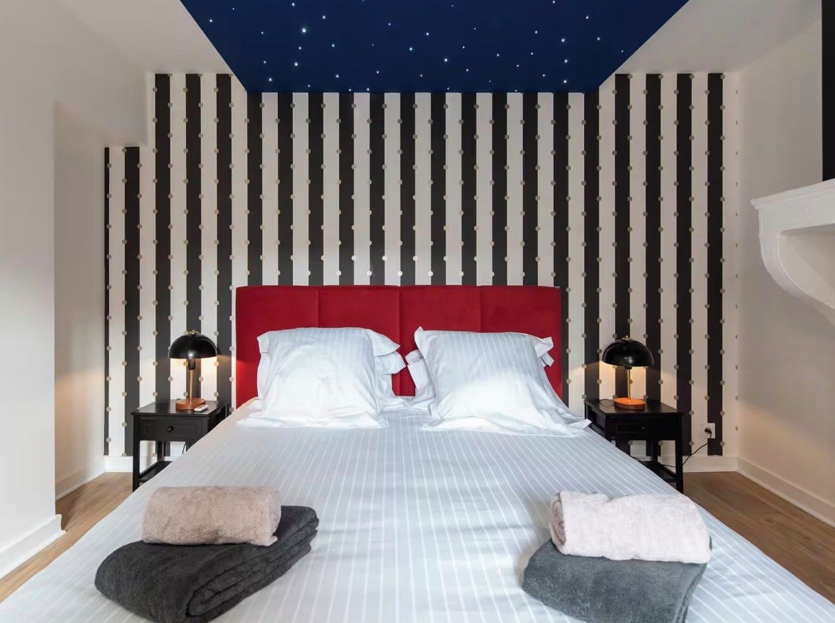 Una habitación de hotel con techo estrellado, cabecera roja, dos mesitas de noche con lámparas y una cama bien hecha con almohadas y toallas.