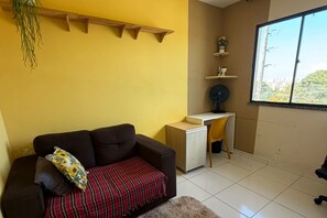 Et værelse med en brun sofa, et lille skrivebord, en stol, et vindue med udsigt og en hylde med planter.