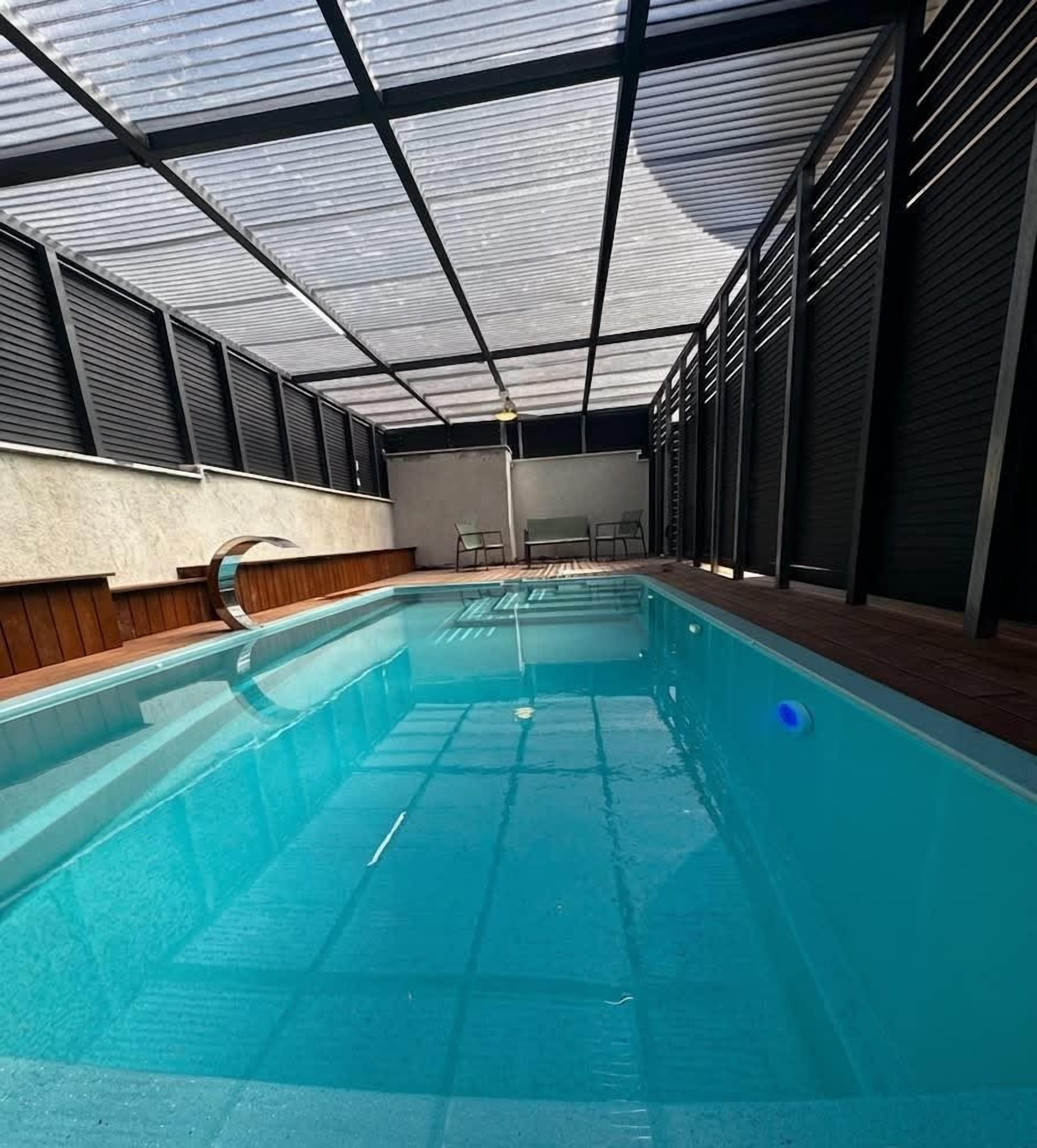 Une piscine intérieure dotée d’un toit en verre, entourée de murs avec des volets métalliques.