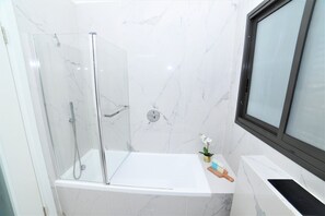 Een badkamer met een douchecabine van glas, een witte badkuip en een raam.