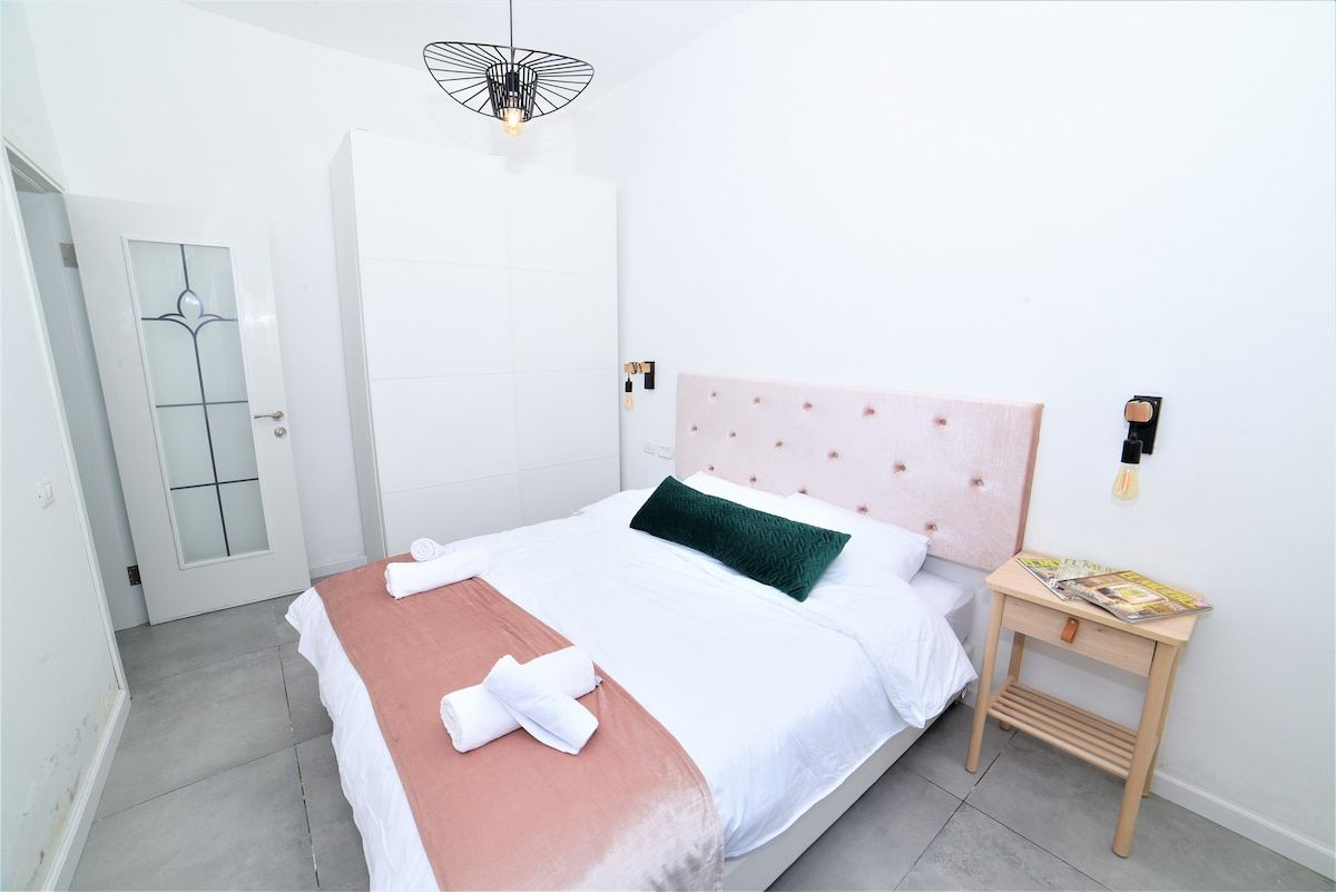 Een net opgemaakt bed met een roze hoofdbord, wit beddengoed en een groen kussen. Er staat een houten nachtkastje met een tijdschrift en een lamp.
