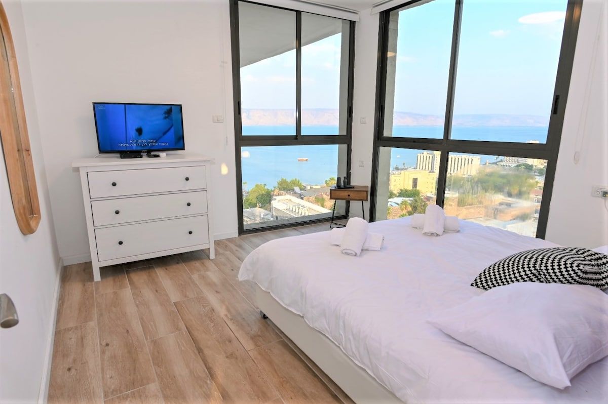 Een slaapkamer met een bed, een televisie op een dressoir en een groot raam met uitzicht op de stad.