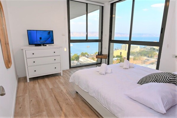 Een slaapkamer met een bed, een televisie op een dressoir en een groot raam met uitzicht op de stad.