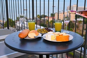 Een tafel met een kaasplank, brood, olijven en twee glaasjes sap op een balkon met uitzicht over de stad.