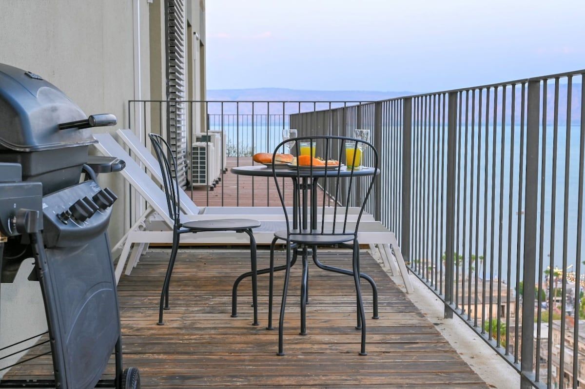 Een balkon met een barbecue, twee stoelen, een tafel en uitzicht op zee.