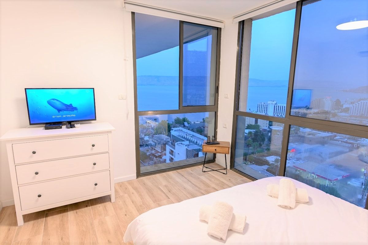 Een hotelkamer met een bed, een televisie op een dressoir en grote ramen met uitzicht op de stad.