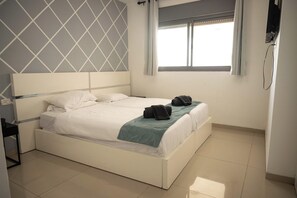 Een nette, moderne hotelkamer met een groot bed, nachtkastje, stopcontact en raam met jaloezieën.