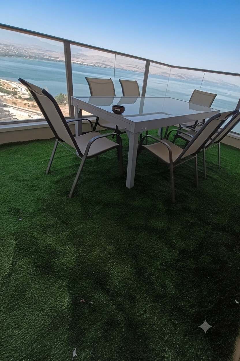 Een tafel met glazen blad en vier stoelen, omringd door een groen gazon, met uitzicht op een waterpartij en een veraf gelegen landschap.
