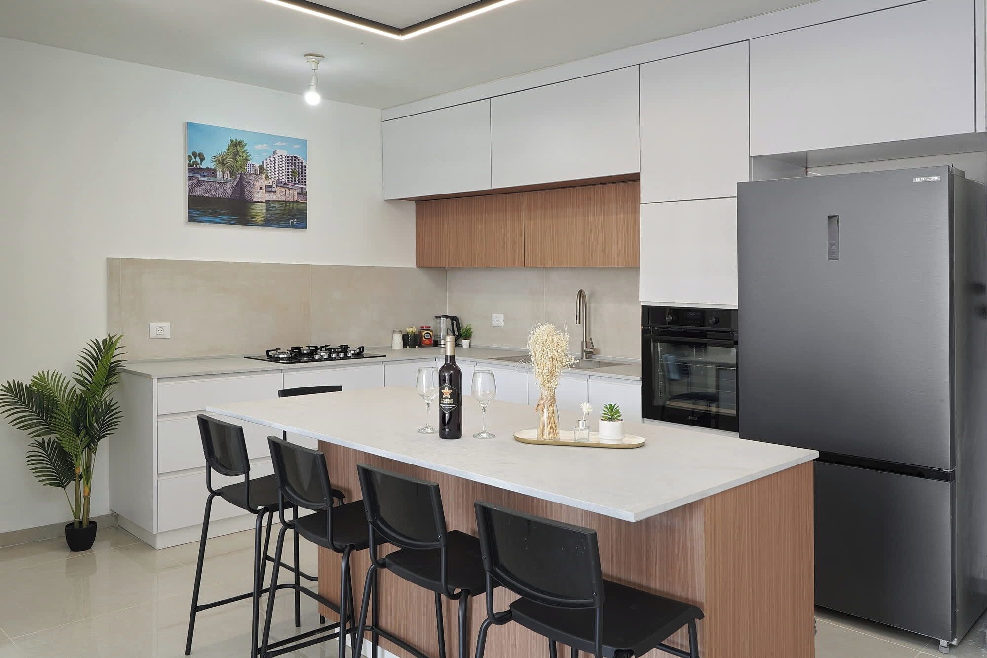 Een moderne keuken met een centraal eiland, barkrukken, een koelkast en een schilderij aan de muur.