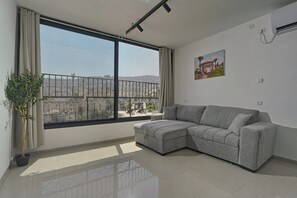 Een moderne woonkamer met een groot raam, een grijze modulaire bank, een potplant en een luchtverkoeler aan de muur.