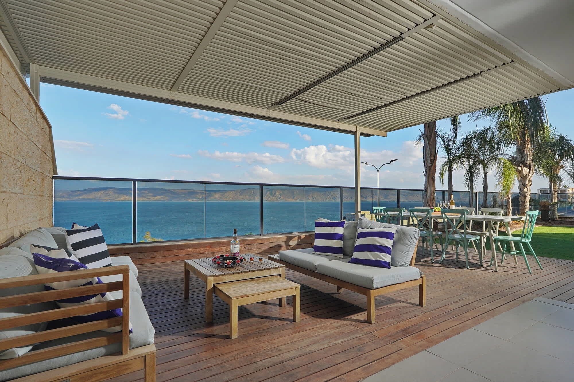 A covered outdoor seating area with a view of the sea, wooden furniture, and a table with a bottle.