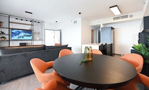 Una sala moderna con un sofá, sillones naranjas, una mesa redonda, un televisor en un estante montado en la pared y una cocina al fondo.