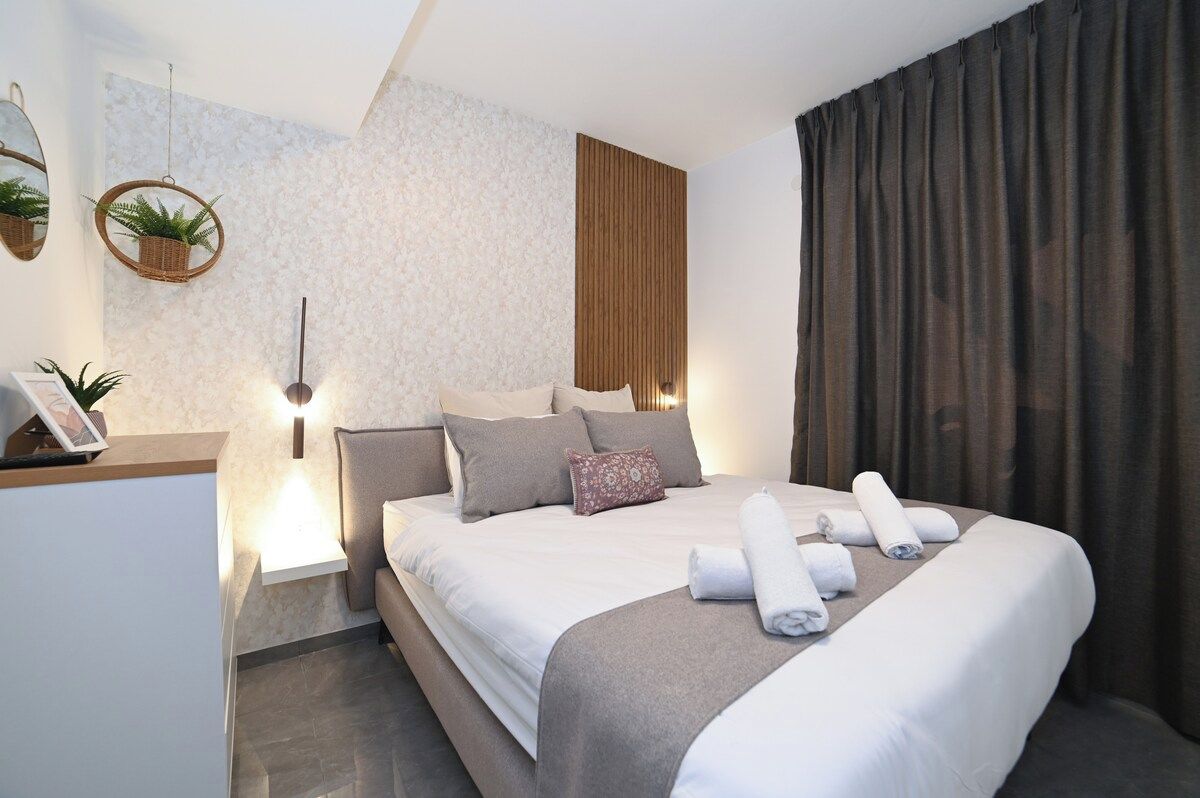 Une chambre d’hôtel moderne, équipée d’un lit, de tables de chevet, d’une lampe fixée au mur et d’une grande fenêtre avec des rideaux.