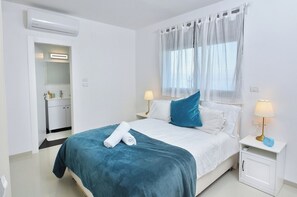 Een hotelkamer met een bed, nachtkastje met lamp, raam met dunne gordijnen en een badkamer met spiegel en wastafel.