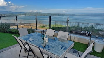 Een terras met een glazen tafel en stoelen, met uitzicht op een waterpartij en heuvels in de verte.