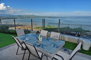 Een terras met een glazen tafel en stoelen, met uitzicht op een waterpartij en heuvels in de verte.