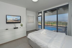 Une chambre d’hôtel équipée d’un lit, d’une télévision, d’une commode et d’une grande fenêtre donnant sur une piscine et un paysage verdoyant.