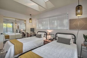 Una camera d'albergo con due letti, un ampio specchio, una finestra con tende, un lampadario geometrico a parete e un tavolino con una pianta.