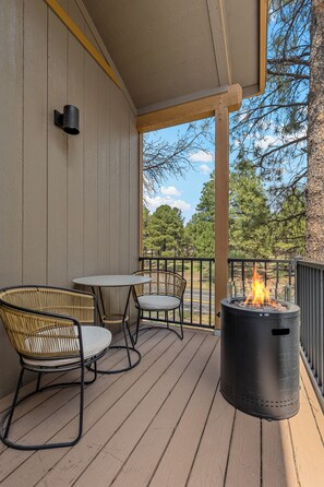 A wooden deck with a round table and two wicker chairs, a black fire pit, and a view of trees and a road. - Central 2BD/2.5Bth - Fire pit , Fast Wifi, Sleeps 5 (Flagstaff)