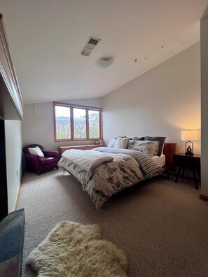 Ein Schlafzimmer mit einem großen Bett, einem Sessel, einem Schreibtisch, einer Lampe und einem Fenster mit Blick.
