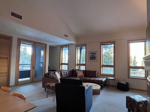 Ein geräumiges Wohnzimmer mit einer großen Eckcouch, einem Couchtisch und einem Esstisch. Große Fenster sorgen für natürliches Licht und Ausblick ins Freie.