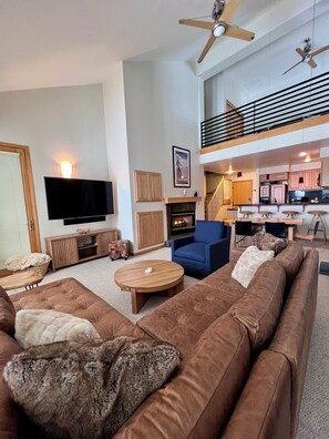 Un salon moderne avec un grand canapé d’angle marron, un fauteuil bleu et une table basse en bois. Un téléviseur à écran plat est fixé au mur, il y a une cheminée et un escalier menant à un niveau supérieur.