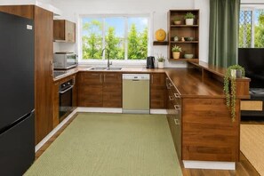 A modern kitchen with wooden cabinets, a dishwasher, and a window with a view of greenery. - Arty Beach Shack Deception Bay | Pets + Parking (Deception Bay)