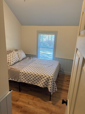 A small, single-bed room with a window, wooden flooring, and a door. - Delightful 2-bedroom cabin in Vanderbilt perfect for a getaway (Vanderbilt)