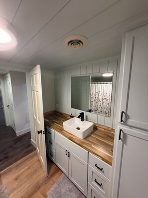 A bathroom with a white sink, wooden countertop, and a mirror above it. There are cabinets with black handles and a door leading to another room. - Delightful 2-bedroom cabin in Vanderbilt perfect for a getaway (Vanderbilt)