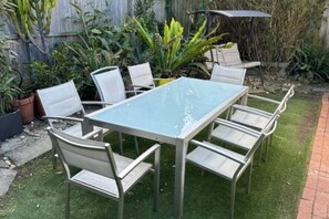 Mesa rectangular al aire libre con cuatro sillas, rodeada de plantas en macetas y una valla de madera.