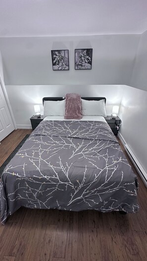 A bedroom with a bed, two bedside tables, a nightstand, a wall-mounted lamp, and two framed pictures on the wall. - Creekside Haven Suite (Moncton)
