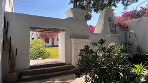 Edificio de paredes blancas con una escalera de piedra que conduce a una puerta, rodeado de follaje y plantas con flores.