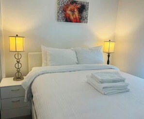 A hotel room with a bed, two bedside lamps, a nightstand, and a painting on the wall. - DOWNTOWN LAKEVIEW LUXE 2BD apartment in marvelous Kelowna with AC and comfort (Kelowna)