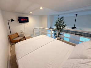 Quarto com cama, escrivaninha, cadeira, planta, TV exibindo 'NETFLIX' e varanda com vista.