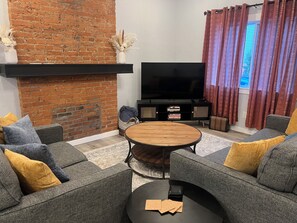 Una sala de estar con pared de ladrillo, un televisor de pantalla plana, sofás grises con cojines amarillos, una mesa de centro redonda de madera y una estantería.