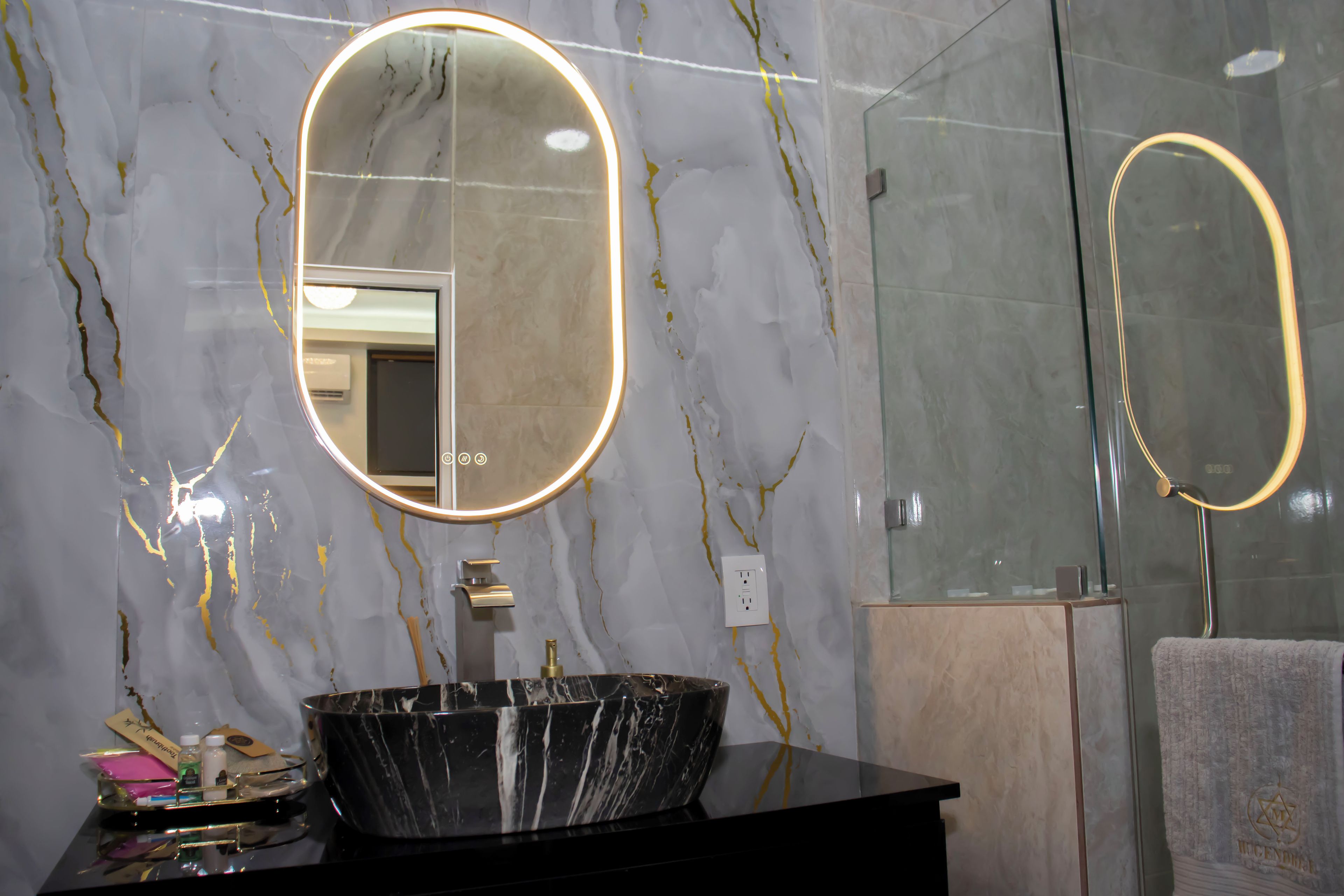 A bathroom with a black marble sink, a round mirror with a light, and a glass shower enclosure.