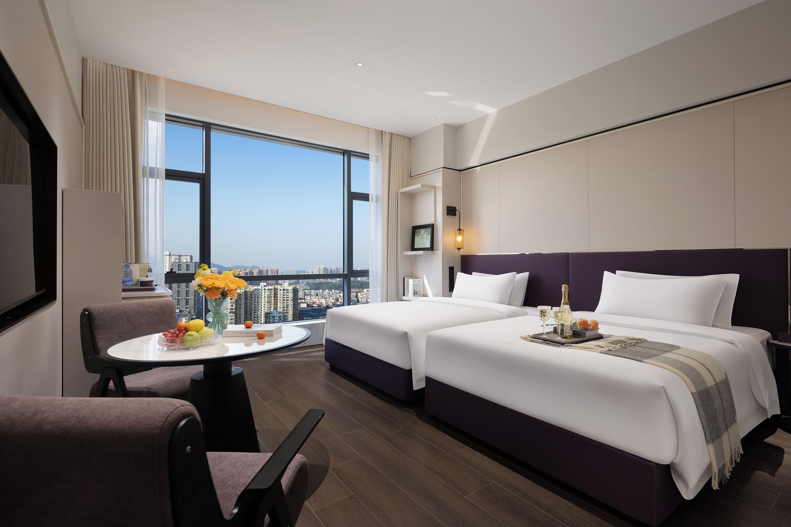 A hotel room with two beds, a dining table with fruit, a view of the city, and a large window.