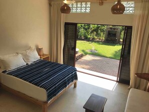 A bedroom with a bed, a nightstand, a small table, and a view of a garden through a glass door. - Private Room Steps to Beach Tropical Garden Retreat Breakfast Included (Punta Perula)