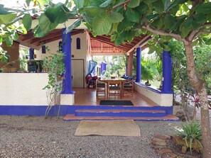 A covered outdoor area with a wooden table and chairs, a blue and white building, and a tree. - Private Room Steps to Beach Tropical Garden Retreat Breakfast Included (Punta Perula)