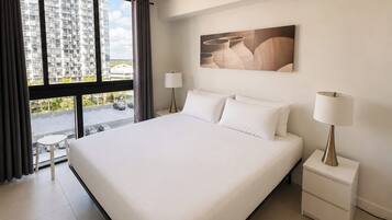 A hotel room with a large bed, two bedside tables, a window with curtains, and a view of a parking lot and buildings.
