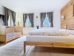 Un dormitorio con cama de madera, cuna, ventana con cortinas, lámpara y un jarrón pequeño con flores.