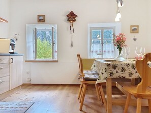Un comedor con una mesa dispuesta para dos, una silla de madera, un reloj en la pared y un ventanal con vistas a árboles.
