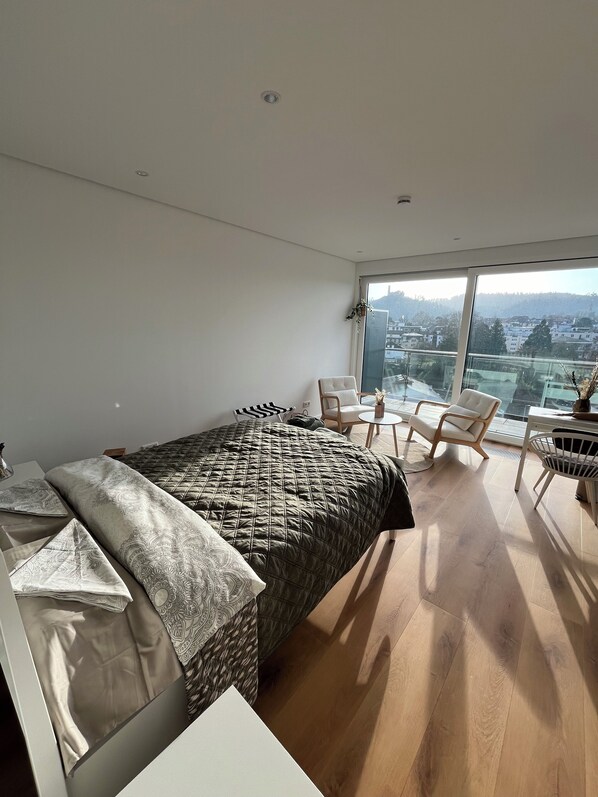 Ein modernes Schlafzimmer mit einem großen Bett, einem Schreibtisch und einem Sessel. Durch große Fenster ist ein Blick auf die Stadt möglich.