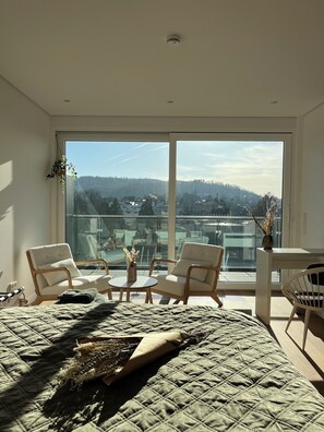 Ein modernes Schlafzimmer mit großem Fenster, zwei Stühlen, einem Bett mit Tagesdecke und einem kleinen Tisch mit einer Vase.