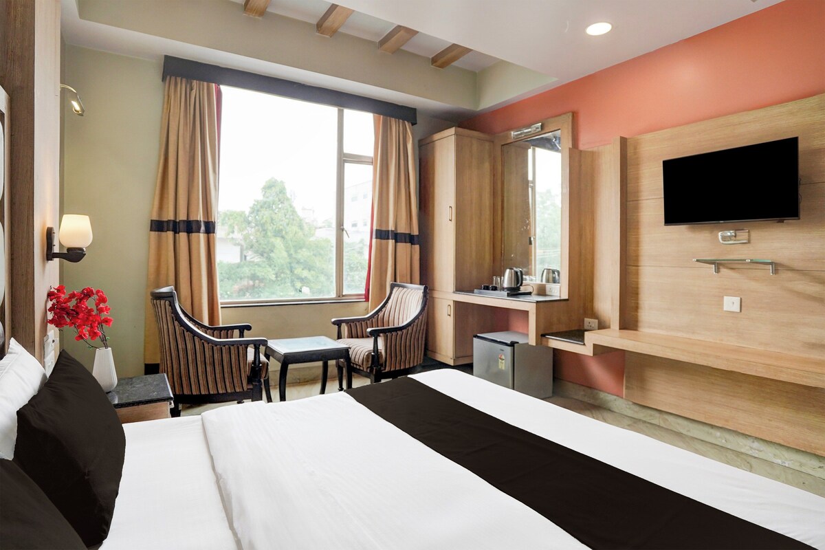 Quarto de hotel com cama, escrivaninha, cadeira, televisão e janela com cortinas.