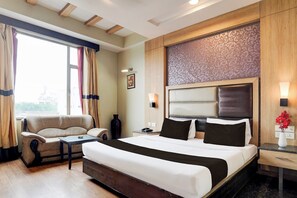 Quarto de hotel com cama, sofá, mesinha, vaso de flores e janela com cortinas.