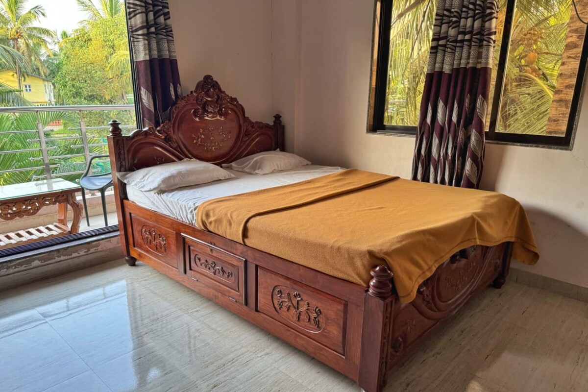 A wooden bed with intricate carvings, a balcony with a view of greenery, and a large window with curtains.