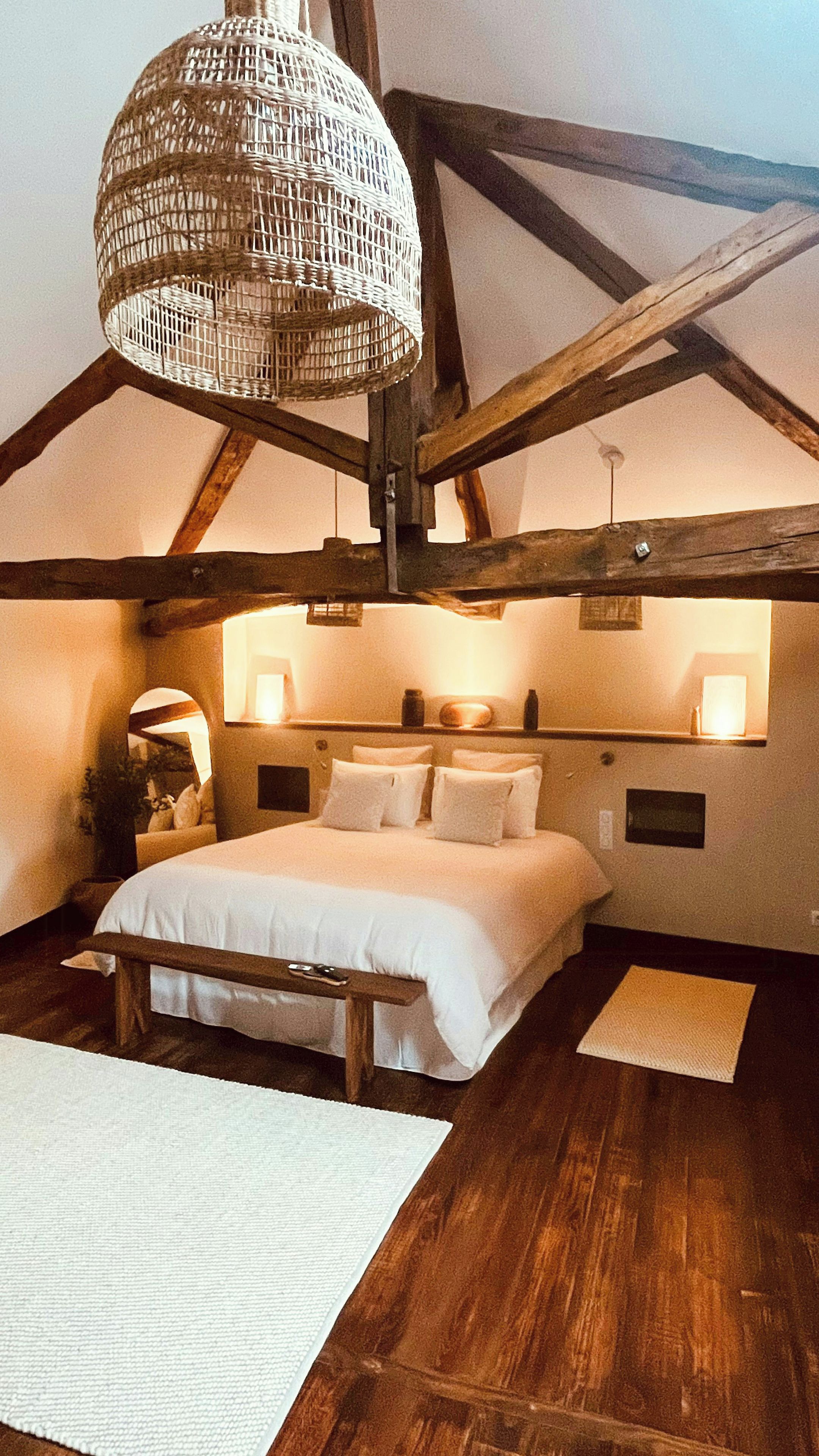 A bedroom with a large bed, wooden beams, a hanging light fixture, and a bedside table.