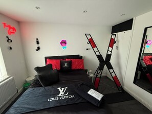 Une chambre avec une couvre-lit noire, des oreillers rouges, une échelle noire et rouge, un panneau lumineux et un miroir.